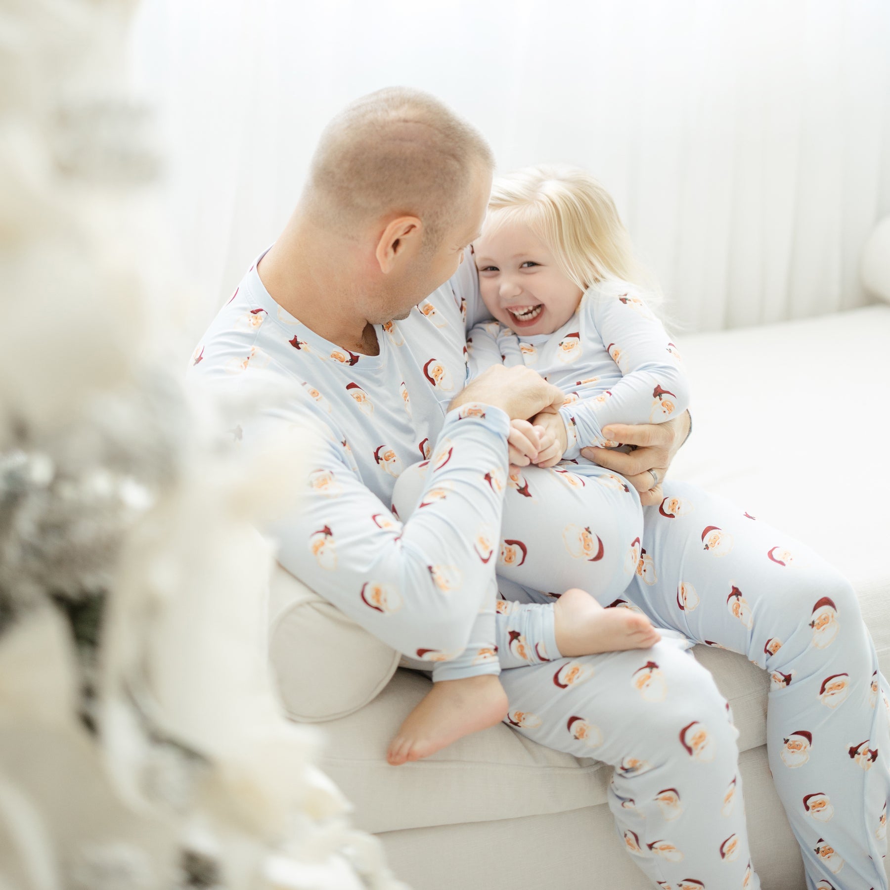 Father sitting on a couch wearing the Men's Jogger Set in Vintage Santa tickling his daughter on his lap who is wearing matching long sleeve toddler pajamas