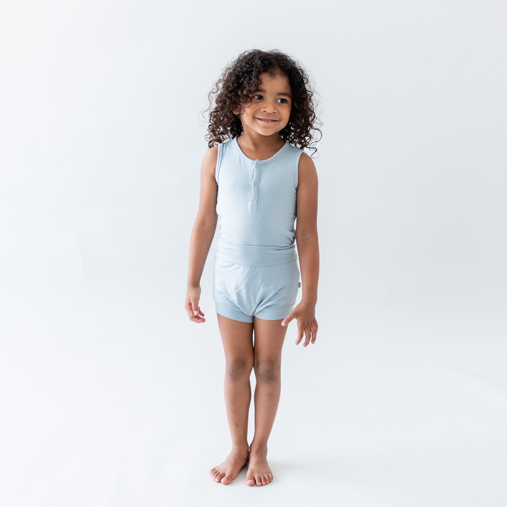 Toddler standing in a white room wearing Kyte Baby Bummies in Fog and matching bodysuit 
