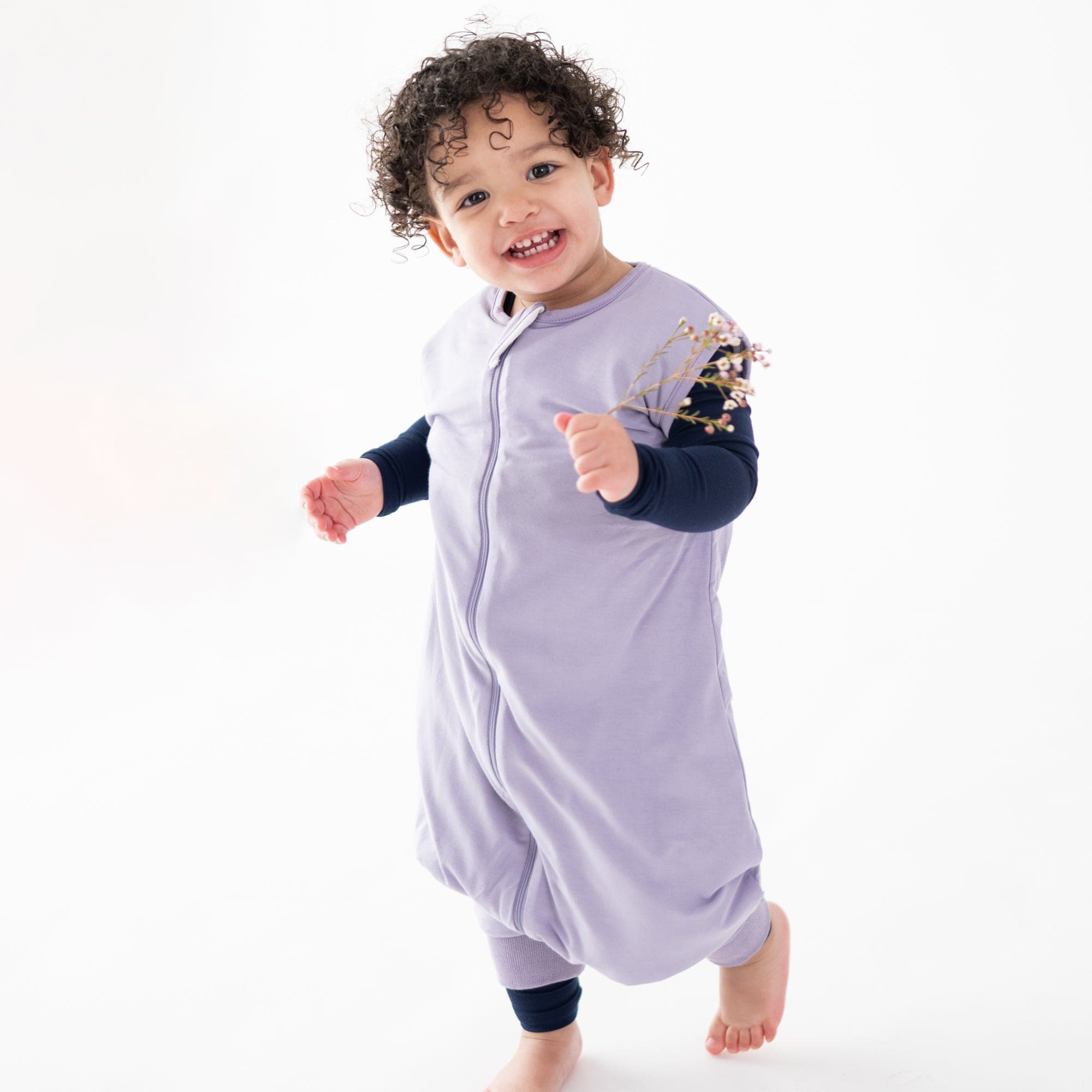 Toddler wearing Kyte Baby Sleep Bag Walker in Taro 1.0 holding flowers