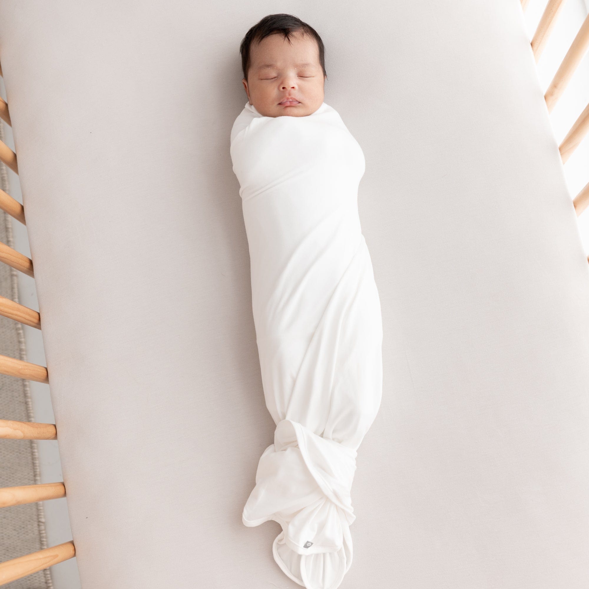 Sleeping newborn laying on Kyte Baby in Oat crib sheet while swaddled in Kyte Baby Infant Swaddle Blanket in Cloud